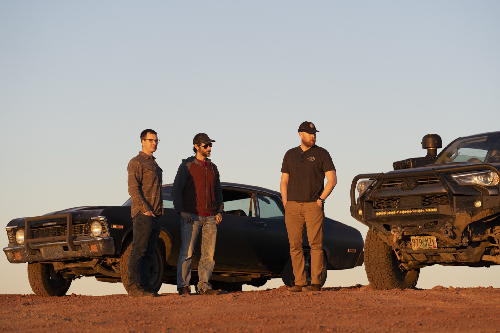 Three crew members with a matte-black muscle car and truck in the desert