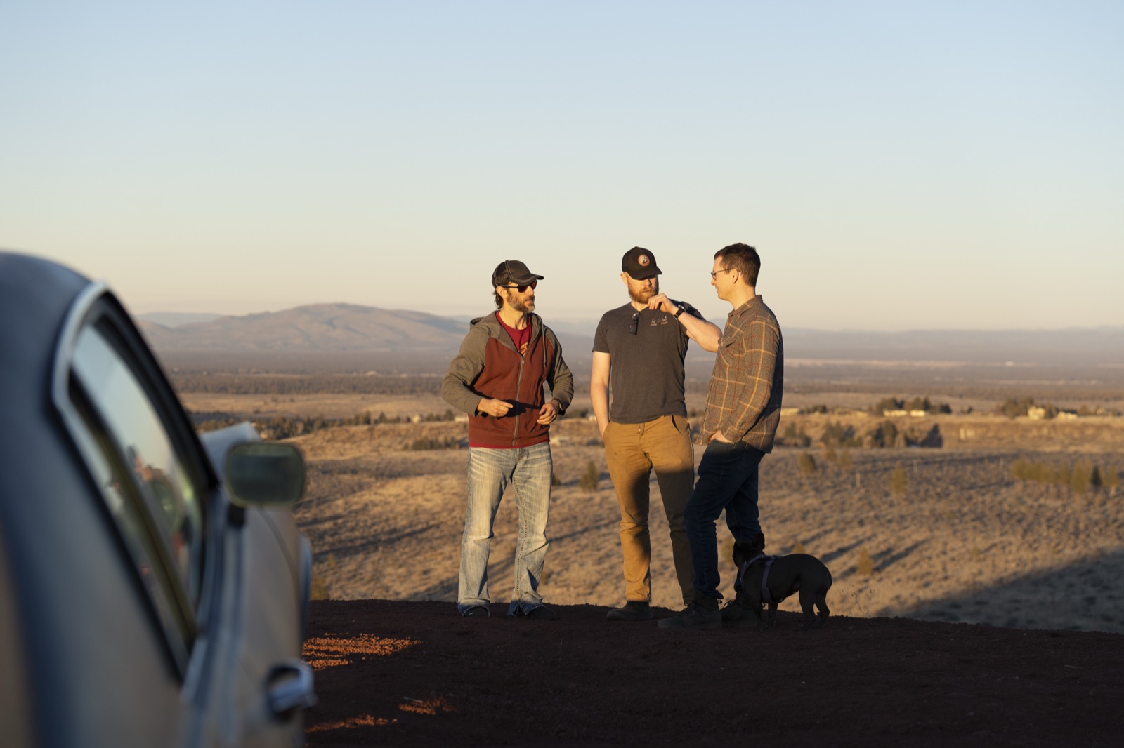 Three crew members and a dog beside a classic car at golden hour