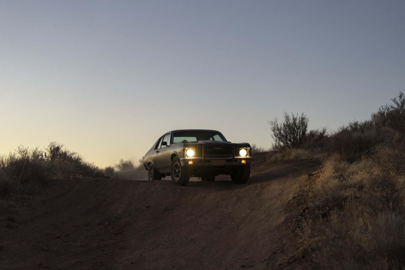 A matte-black muscle car cresting a dirt road at dusk, headlights on