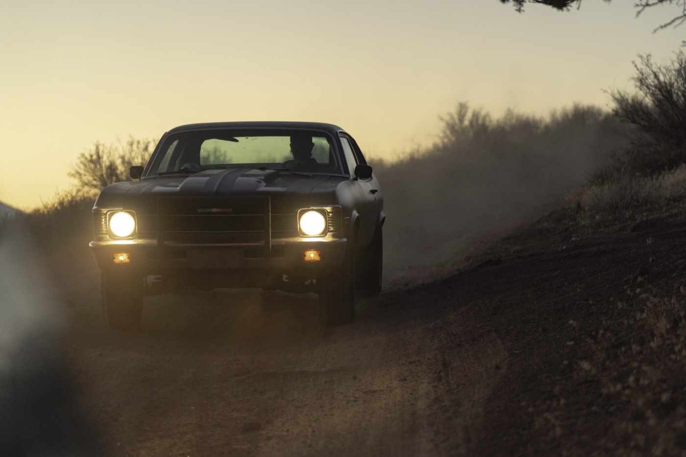 The car head-on at dusk, headlights cutting through the dust